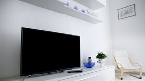 Un salon moderne avec un téléviseur à écran plat posé sur un meuble blanc, un bol bleu, une chaise blanche et une étagère ornée d’objets décoratifs.