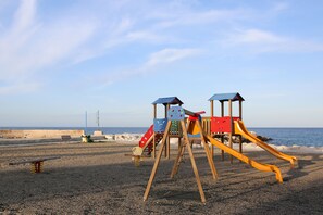 Un parque infantil con toboganes y columpios en una playa de guijarros, con el mar al fondo.
