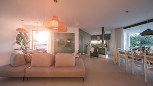 Ein modernes Wohnzimmer mit einem beigen Sofa, einem Essbereich mit Holztisch und -stühlen sowie großen Fenstern, die viel Tageslicht hereinlassen.