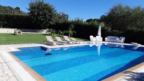 A rectangular swimming pool with a tiled edge, surrounded by a stone border. There are white lounge chairs and a white umbrella on the poolside.