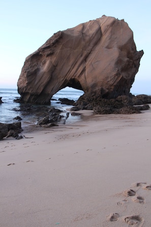 A large rock formation with an arch over the ocean, sandy beach, and visible footprints. - Casa Milvus - Between the beach and the countryside (São Pedro da Cadeira)