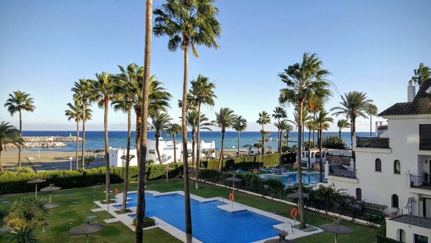 Área da piscina com palmeiras, vista clara do oceano e um prédio com varandas.