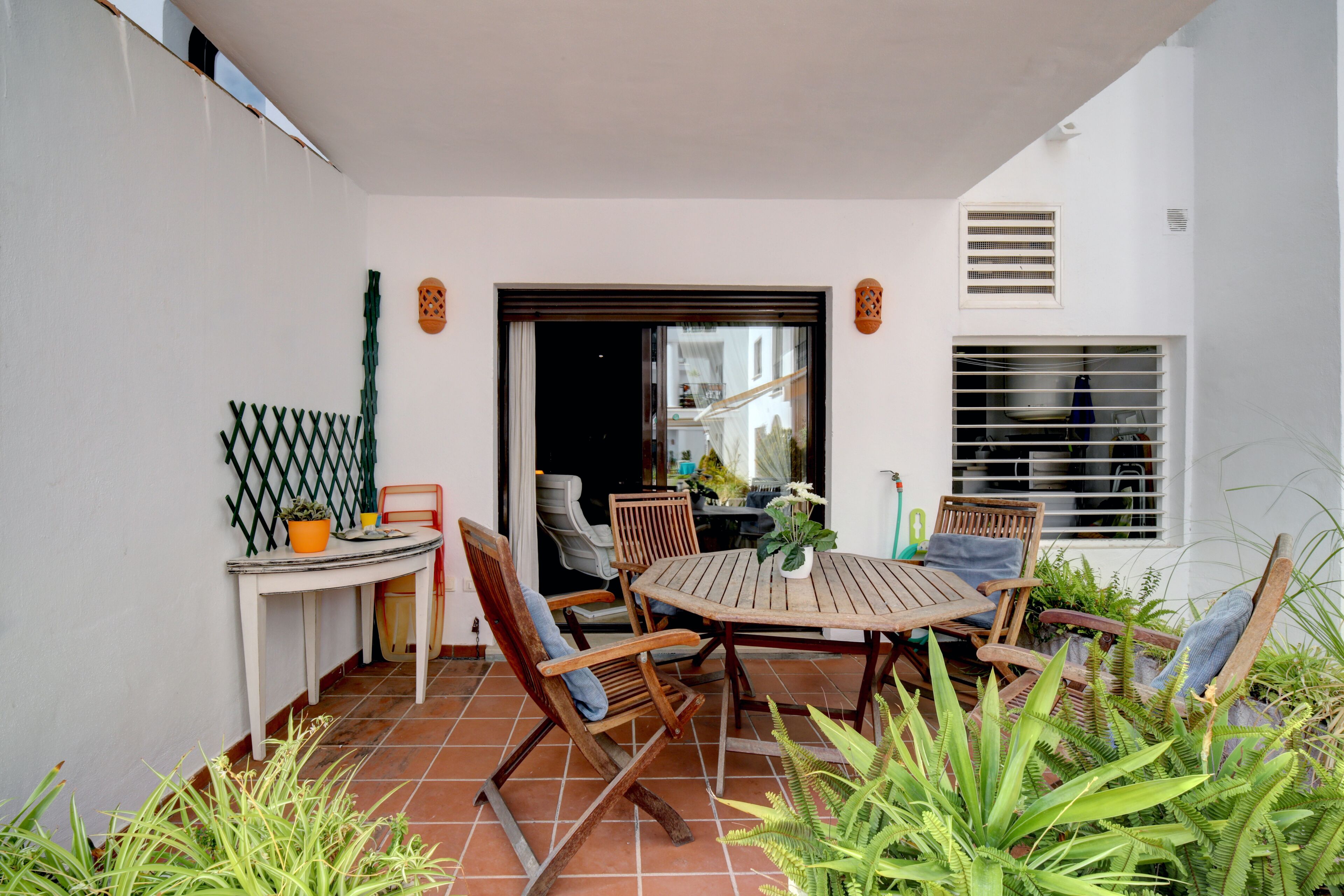 A patio with a round wooden table and chairs, surrounded by potted plants and a white wall.
