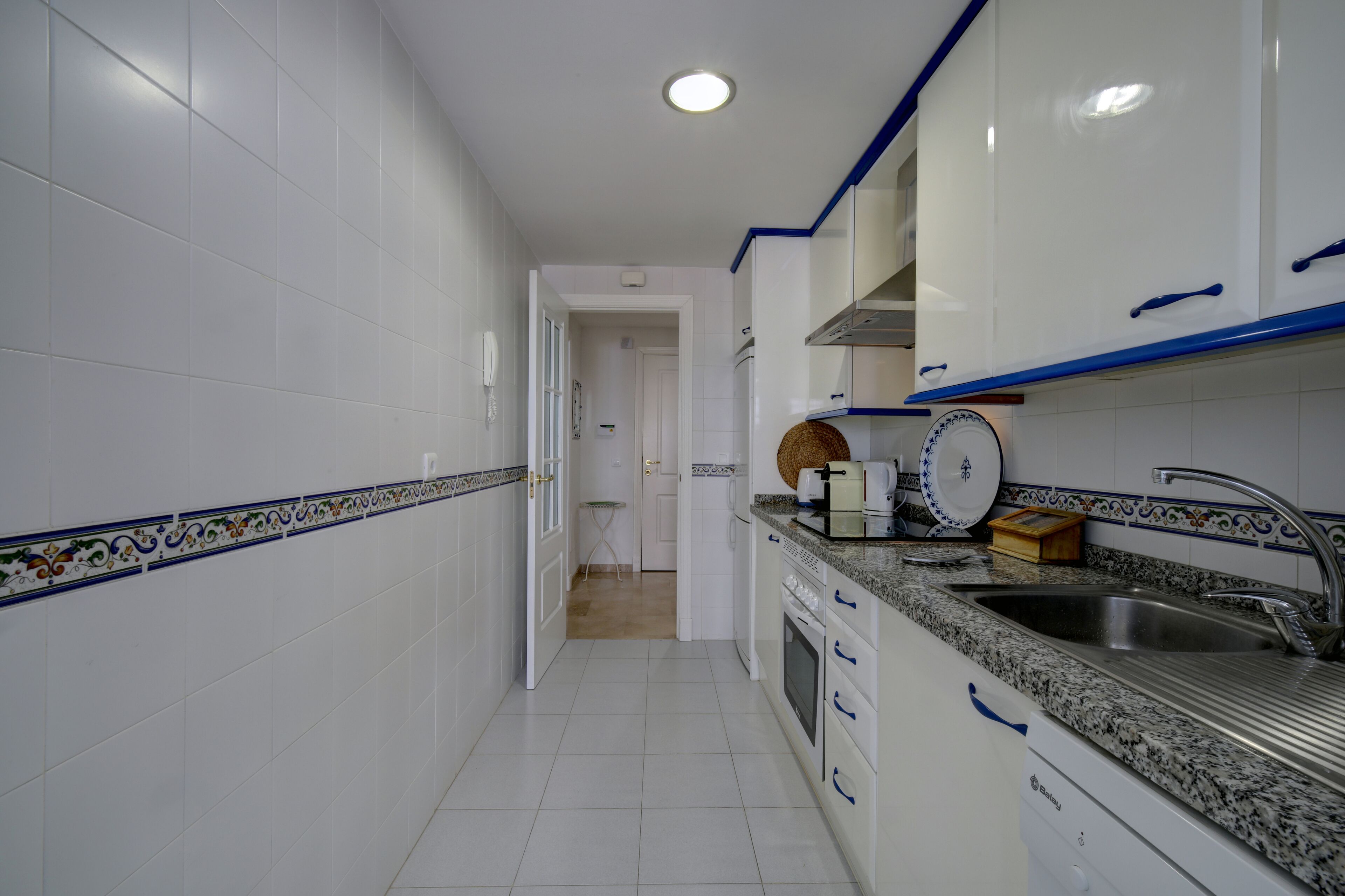 A narrow kitchen with white cabinets, a granite countertop, and a stainless steel sink.