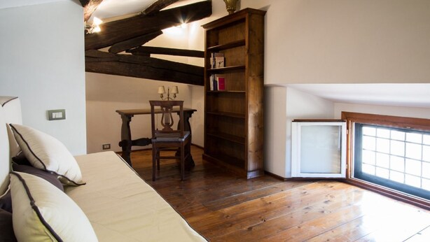 A room with a wooden floor, a bed with pillows, a desk with a lamp, a bookshelf, and a window with blinds. - ALTIERI LOFT PANTHEON (Roma)