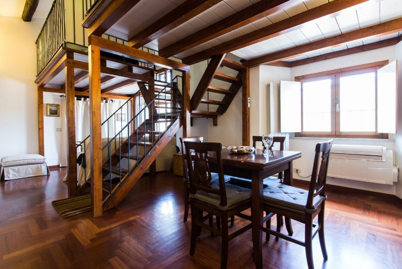 Una habitación amplia con una escalera de madera, una mesa de comedor con sillas y un ventanal que proporciona luz natural.