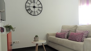 Sala de estar con sofá beige, cojines estampados, una mesita auxiliar, una planta en maceta y un reloj de pared grande.