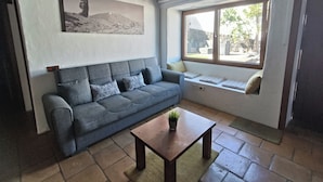 Ein Wohnzimmer mit grauem Sofa, einem Holztisch, einem Fenster mit Aussicht und einem Bild an der Wand.