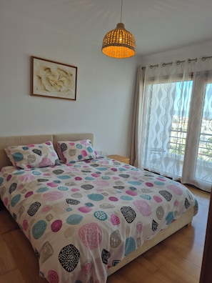 A bedroom with a bed, a hanging light fixture, a window with curtains, and a painting on the wall.