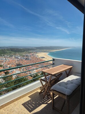 A balcony with a table and chair, overlooking a coastal cityscape with a beach and hilly terrain.