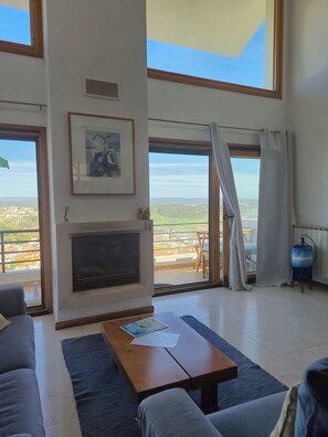 A spacious living room with a fireplace, large windows, and a balcony with a view of the city.