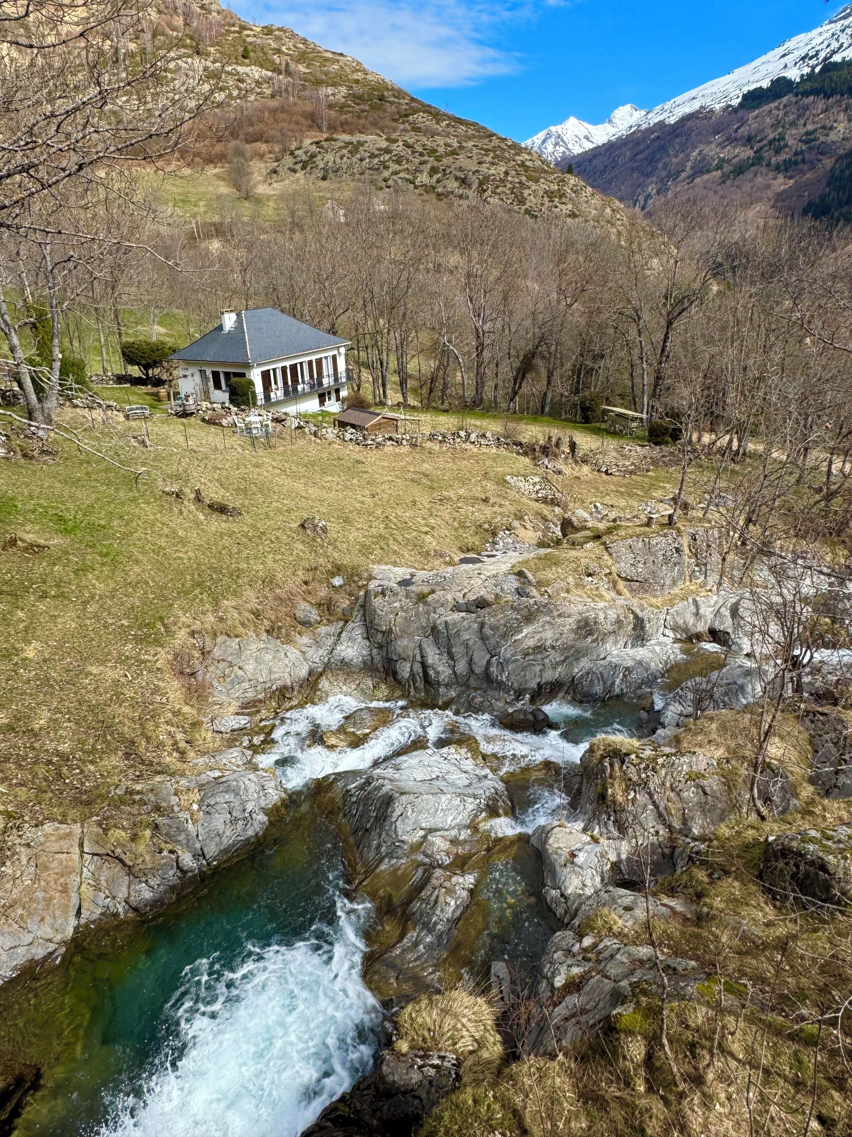 Una casa con balcón, un arroyo y montañas al fondo.