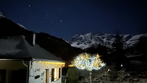 Una casa con terraza de madera y un pequeño jardín, rodeada de montañas nevadas.