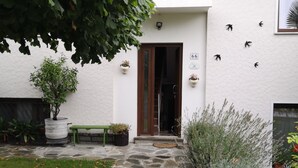 Un sentiero in pietra conduce a un edificio bianco con una porta in legno. Piante in vaso e una panchina si trovano all'esterno.