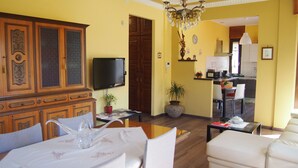 Un soggiorno con televisore, una zona pranzo con tavolo e sedie, una cucina sullo sfondo e un lampadario.
