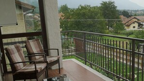Un balcone con mobili in vimini, un tavolo in vetro e vista su una zona residenziale con case e montagne sullo sfondo.