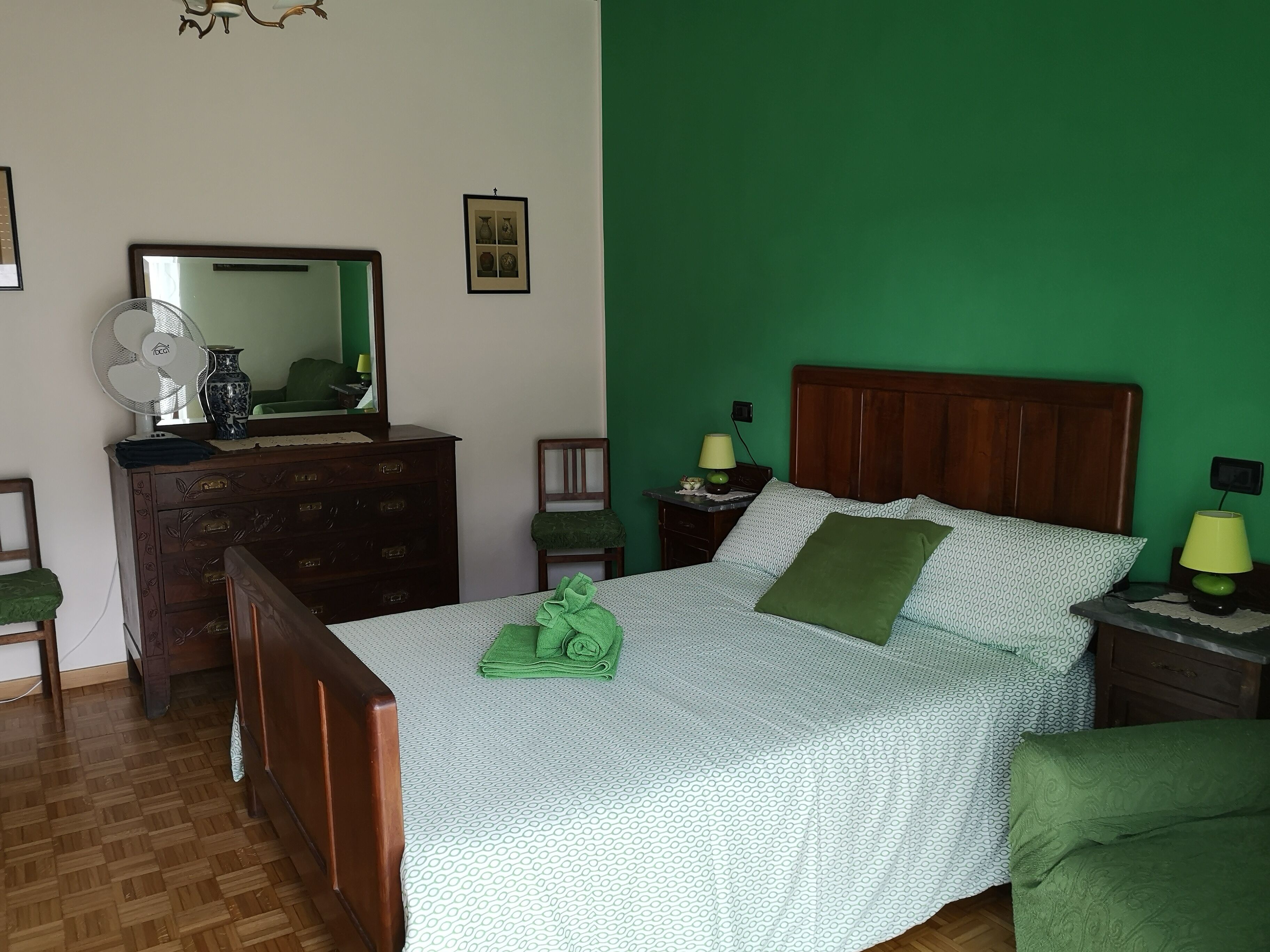 Un dormitorio con cama de madera, paredes verdes, un aparador, un espejo y un ventilador.