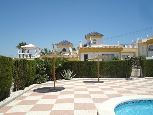 A pool area with a checkered floor, surrounded by a hedge and a building with a yellow facade and a tiled roof.