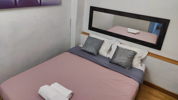 Ein ordentlich bezogenes Bett mit einer rosa Tagesdecke und Kissen, ein großer Spiegel darüber und ein Gemälde eines Piers an der Wand.