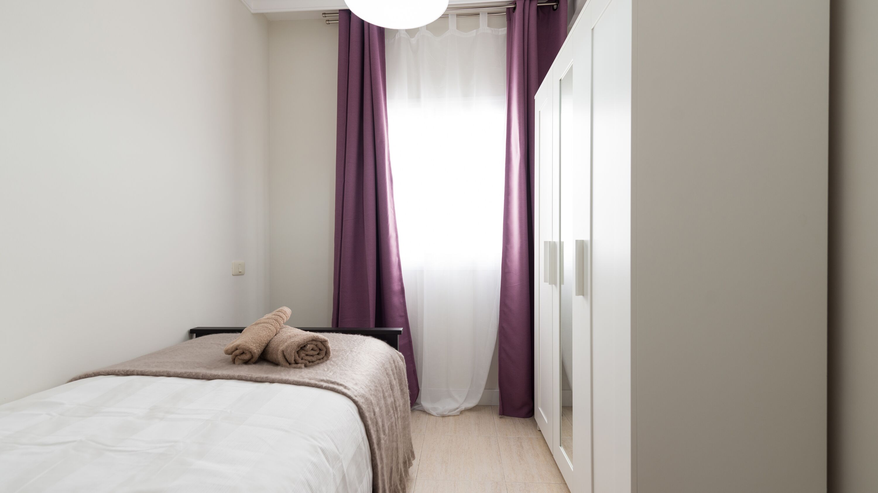 Een minimalistische slaapkamer met een bed, een garderobe en een raam met paarse gordijnen.