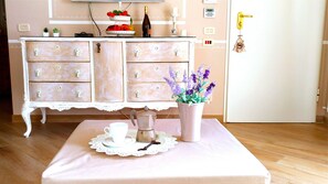 A room with a white dresser, a coffee table with a white tablecloth, a white cup, a French press, and a vase with purple flowers.