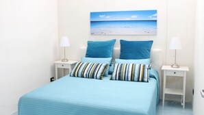 Een slaapkamer met een bed, twee nachtkastjes, een nachtlampje, een schilderij van een strand en een lamp op elk tafeltje.