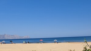 Een strand met mensen die ontspannen onder parasols, een heldere lucht en bergen op de achtergrond.