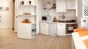 A compact kitchen with white cabinets, a microwave, a dishwasher, and a sink. There are shelves above the counter and a small table with a placemat. - Vatican Luxury Apartment (Rome)