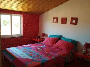 A bedroom with a wooden ceiling, a large window, a bed with colorful bedding, and three wall-mounted picture frames.