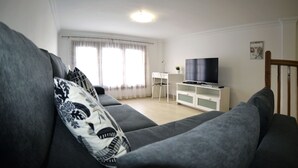Un soggiorno moderno con un divano grigio, una TV a schermo piatto su un mobile bianco, una piccola scrivania e una finestra con tende leggere.
