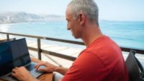 Eine Person benutzt einen Laptop auf einem Balkon mit Blick auf das Meer.