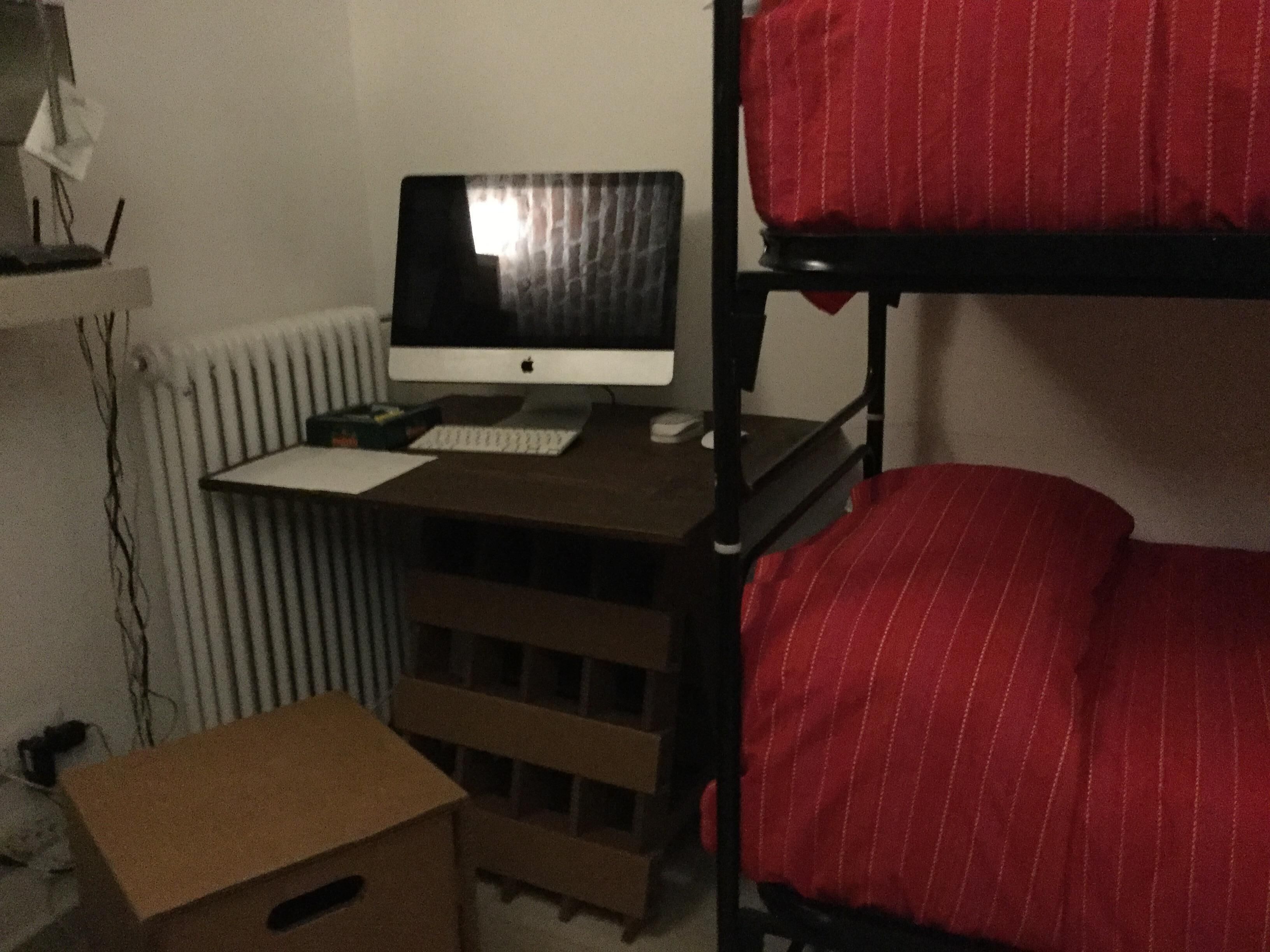 A bunk bed with a red-striped mattress, a desk with a computer, and a cardboard box.