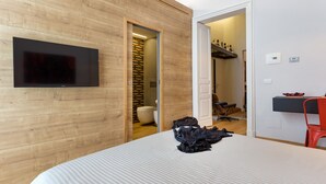 A modern hotel room with a wooden wall, a flat-screen TV mounted above a bed, a red chair, and a desk with a bowl of fruit. - Uflat • comfy apt, in the historic center of Catania. (Catania)