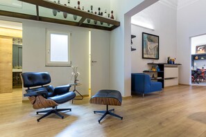 A modern living room with a black leather sofa, a blue armchair, and a wooden ottoman. A glass shelf with decorative items is above the sofa.