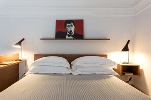 A neatly made bed with white pillows and a striped bedspread, flanked by two bedside tables with lamps, a wooden headboard, and a red framed artwork above.