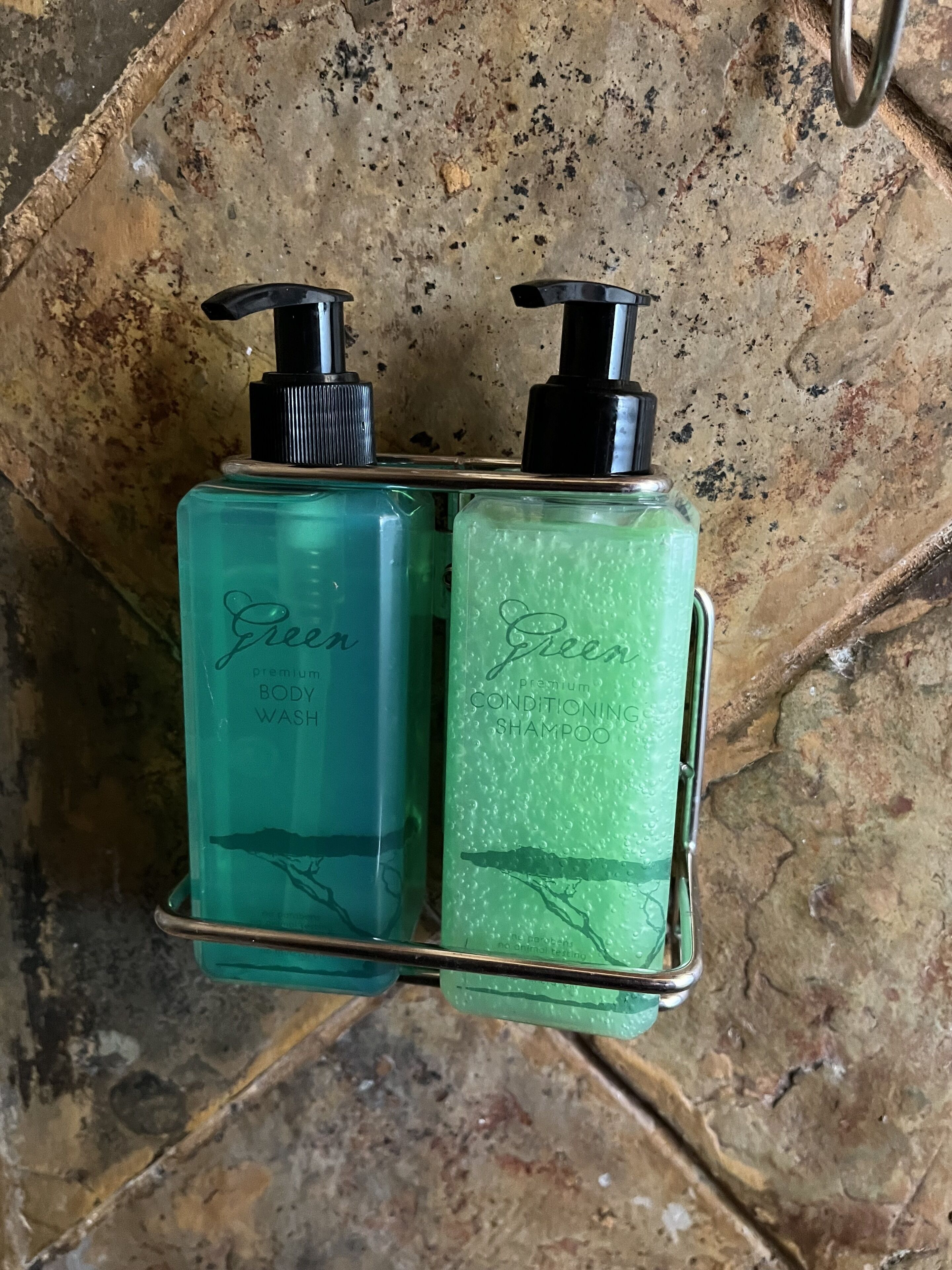 Dois frascos de gel de banho verde e shampoo condicionador em um suporte de metal, diante de uma parede texturizada.