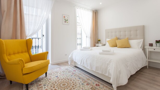 A bedroom with a bed, a yellow armchair, a nightstand, and a window with curtains.