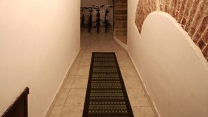 Un couloir étroit avec un sol carrelé, un paillasson noir à l’entrée et un mur de briques sur la droite.