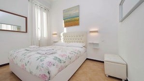 Une chambre à coucher avec un lit, une table de chevet, une commode, un tableau au mur et une fenêtre avec des rideaux.