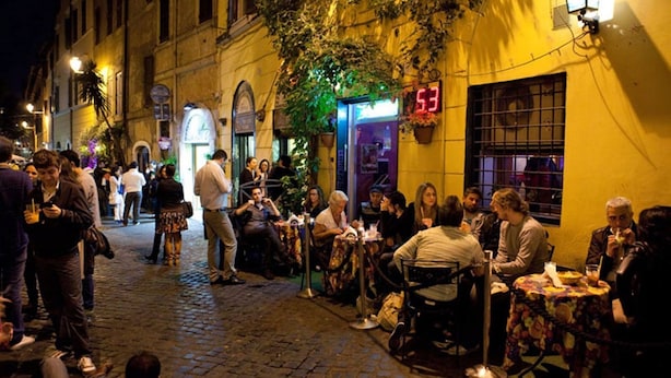 Un café animé en plein air, avec des tables et des chaises, entouré d’immeubles.