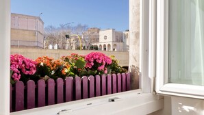 Une fenêtre donnant sur un bâtiment à la façade blanche et orné d’une clôture en bois peinte en violet.