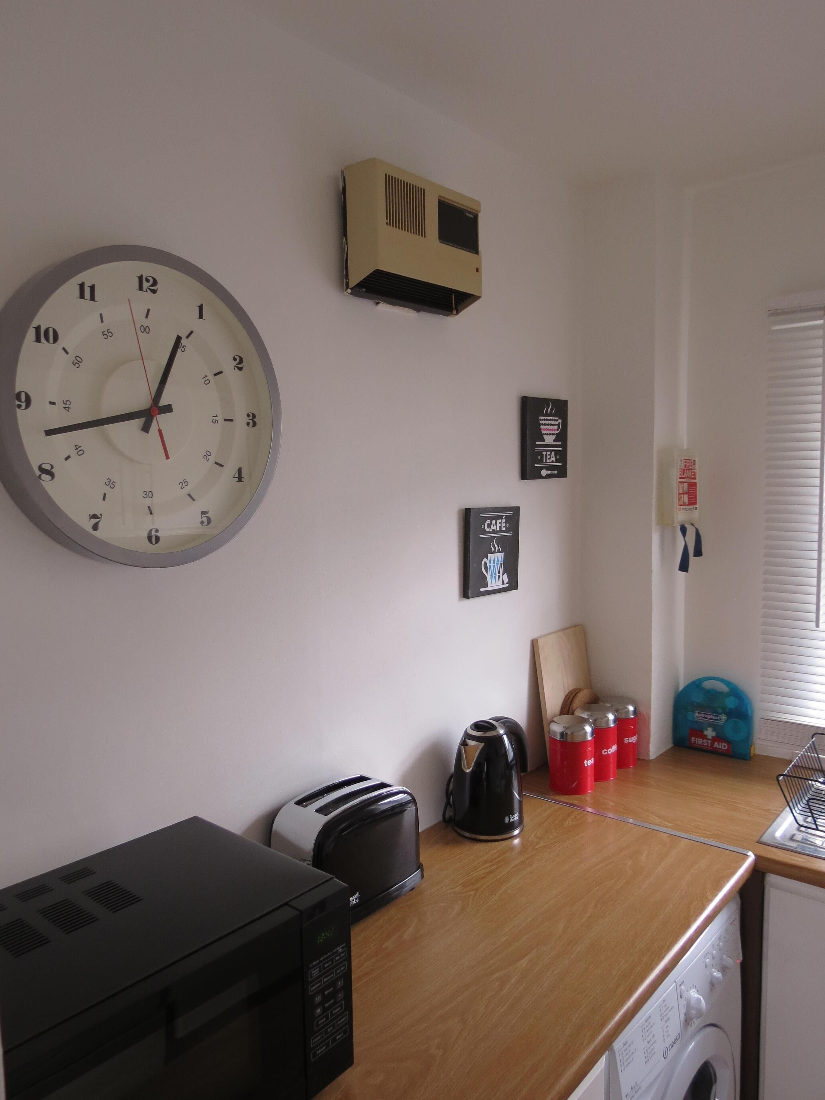 Una cocina con microondas, tostadora, hervidor y cafetera sobre la encimera. Hay un reloj en la pared y un botiquín de primeros auxilios en el suelo.