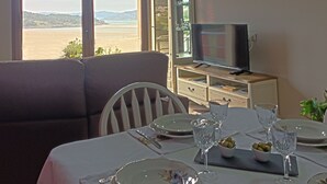 Ein Essbereich mit Tisch für zwei Personen, einer Couch, einem Fernseher auf einer Holzkommode und einem schönen Ausblick durch das Fenster.
