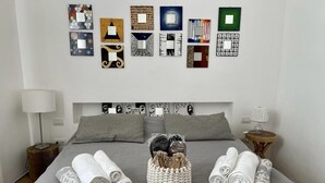 A neatly made bed with grey linens, a woven basket with toiletries, and a wall with various framed artworks. - Bijoux Luxury Apartment in Old Town (Bari)