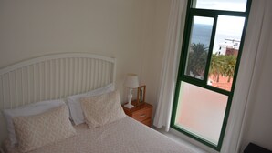 Quarto com cama, mesa de cabeceira, abajur e janela com vista para o mar e edifícios.