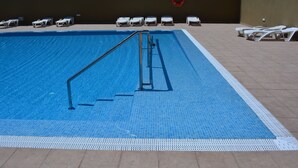 Uma piscina com deck revestido de azulejos, escada de metal e cadeiras de lounge brancas.