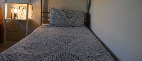 Un llit individual amb un estovat estampat, una taula de nit amb una làmpada i una presa corrent.