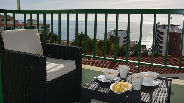 Uma varanda com móveis de vime, uma mesa de jantar com tigelas e xícaras, e vista para o mar e edifícios.