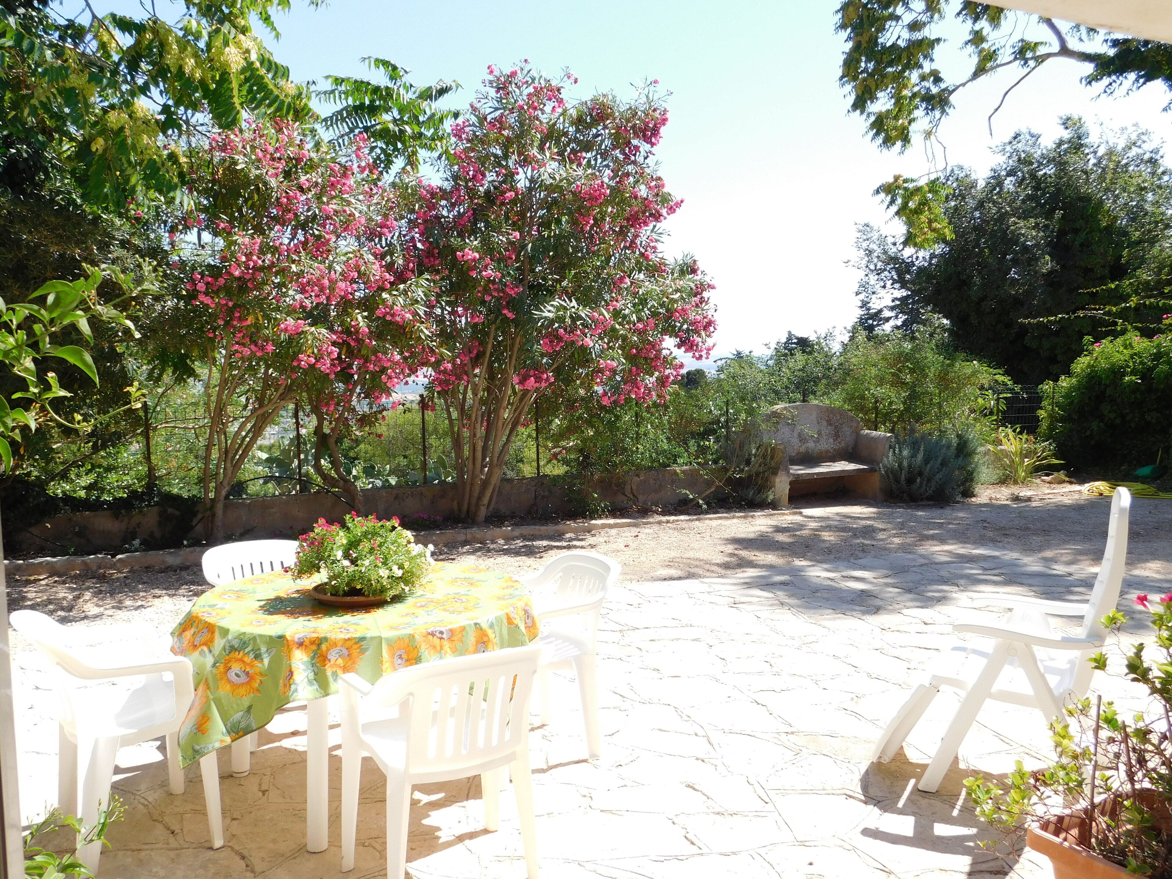 An outdoor patio with a round table and white chairs, surrounded by lush greenery and flowering trees.