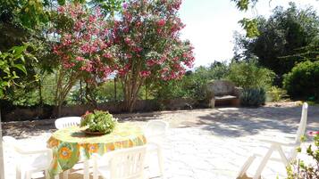 An outdoor patio with a round table and white chairs, surrounded by lush greenery and flowering trees.